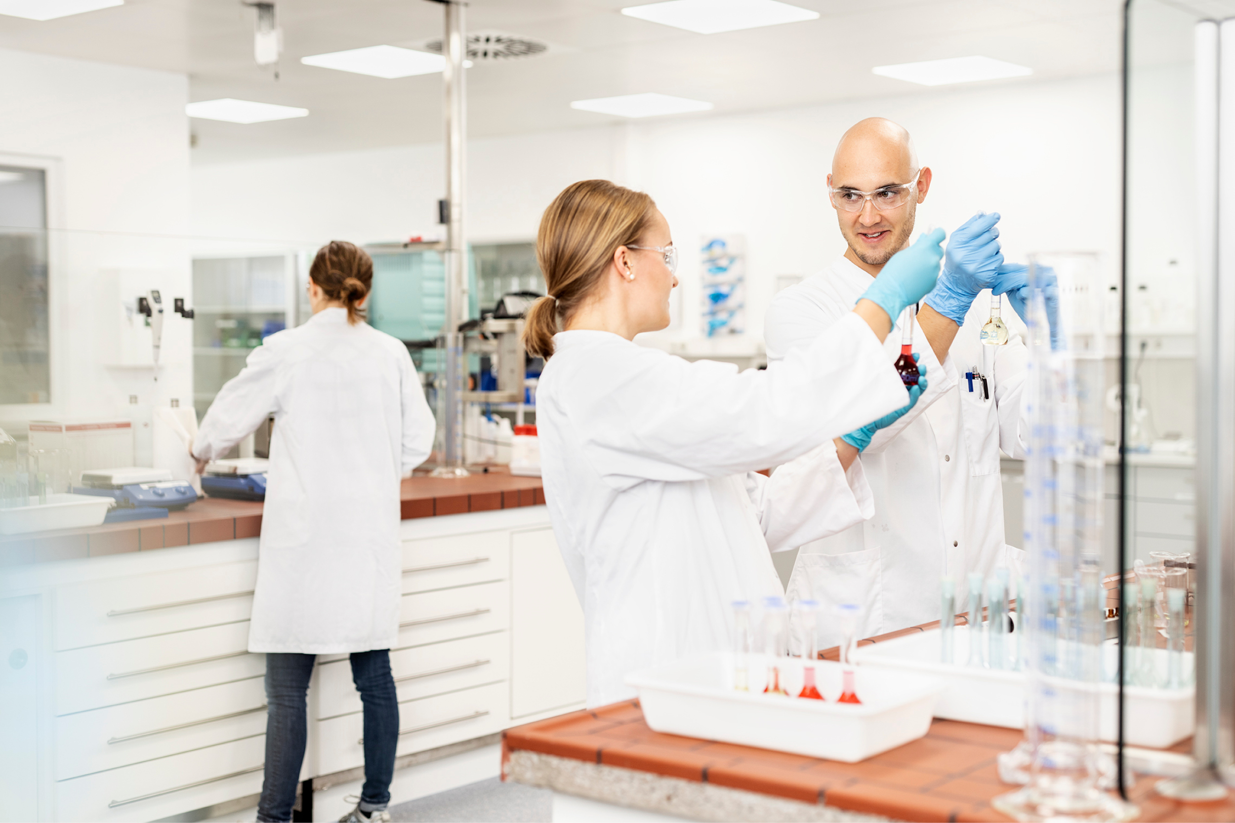 Ein Mann und eine Frau stehen im weißen Kittel in einem Labor und schauen sich an. Im Hintergrund steht eine weitere Person im weißen Kittel.