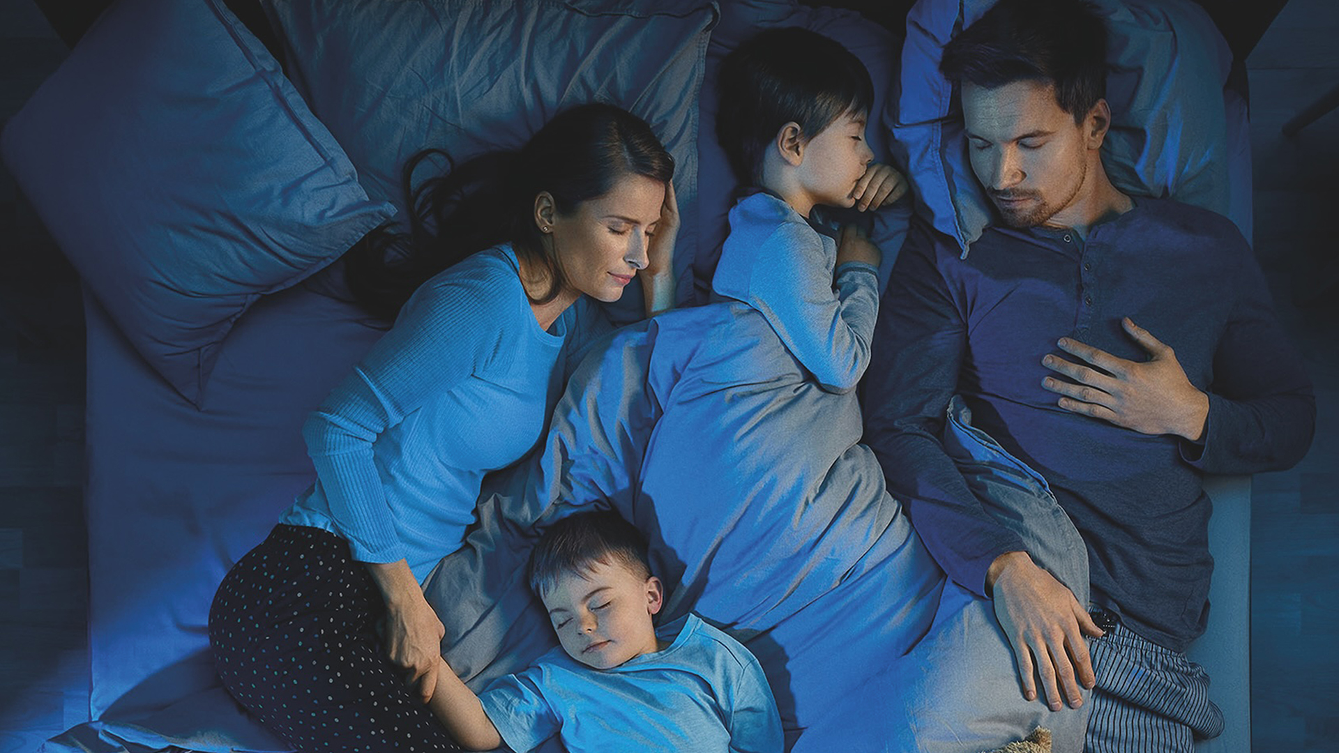 Eine Familie bestehend aus zwei Erwachsenen und zwei Kindern, die friedlich in einem Bett unter bläulichem Licht schlafen.