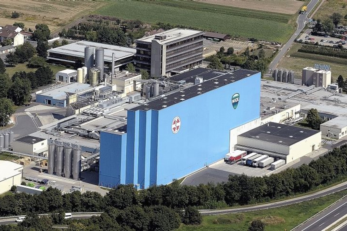 Luftaufnahme des WICK Werks in Groß-Gerau mit großem blauem Produktionsgebäude mit WICK-Logo, angrenzenden Industrieanlagen, Silos und Lagerhallen; umgeben von Bäumen, Feldern und Straßen.